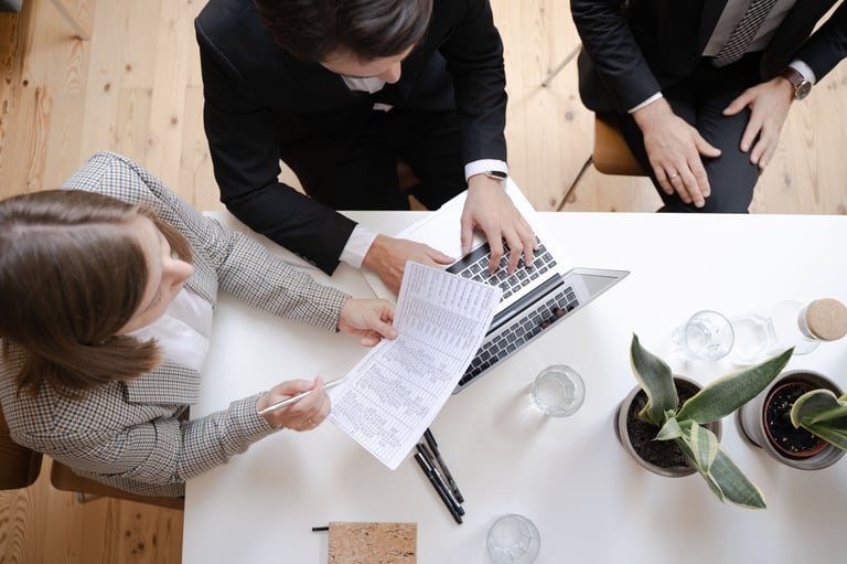 Two businesspeople review documents together at a white table with laptops, coffee cups, and a potted plant