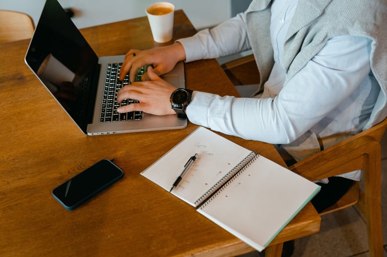 Person in light blue shirt typing on laptop at wooden desk with notebook, pen, phone, and coffee cup